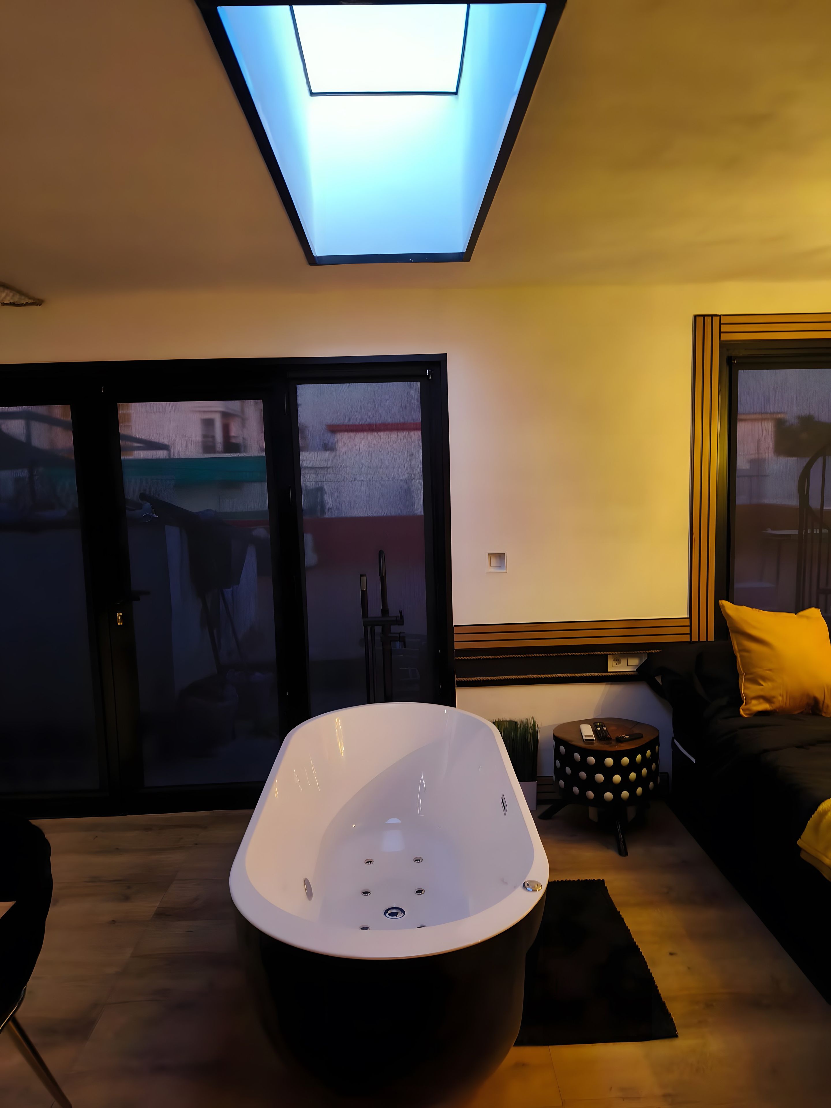 A modern living room with a large bathtub, a sofa with a yellow pillow, and a small round table with a remote control.