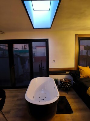 Un salon moderne avec une grande baignoire, un canapé agrémenté d’un coussin jaune et une petite table ronde sur laquelle repose une télécommande.