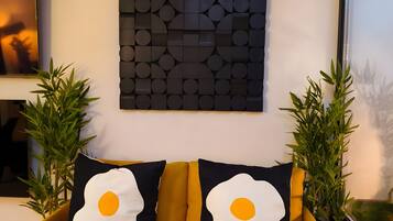 En gul soffa med två svartvita, äggformade kuddar, ett geometriskt konstverk som hänger på väggen och två krukväxter.