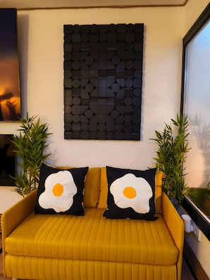 Un sofá amarillo con dos almohadas blancas y negras con forma de huevo, una obra de arte geométrica colgada en la pared y dos plantas en macetas.