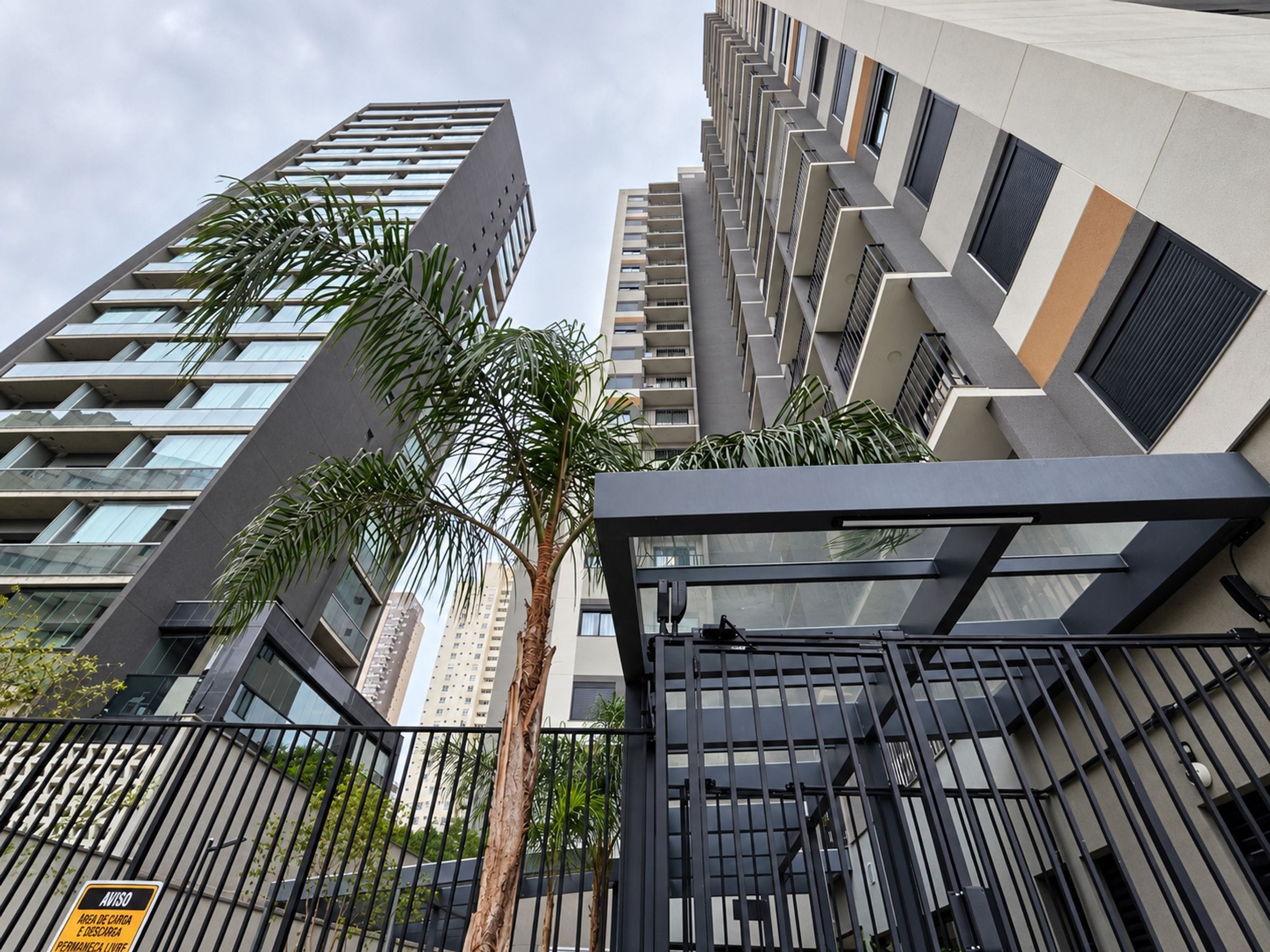 Un immeuble moderne de grande hauteur, doté de nombreux balcons, d’une entrée sécurisée et d’un palmier au premier plan.