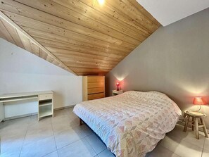 A bedroom with a sloped ceiling, a bed with a patterned bedspread, a bedside table with a lamp, and a desk with a chair. - 3rd floor, view panoramic, terrace, tv, 68m², Serre Chevalier (Briançon)
