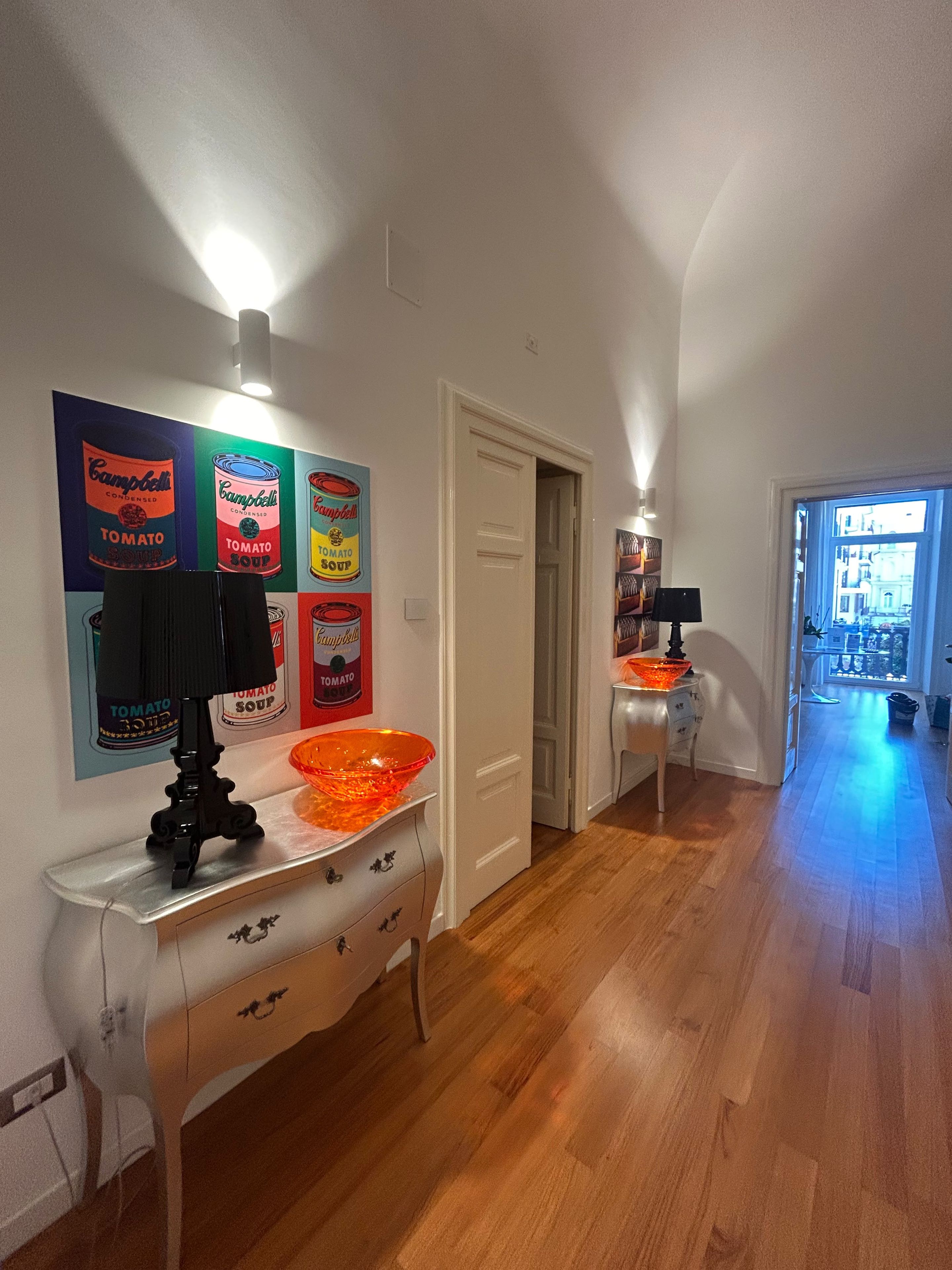Un pasillo con una puerta blanca, una mesa blanca con una lámpara negra y un cartel de colores en la pared.