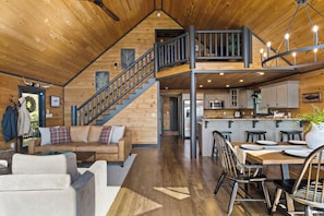 A spacious living area with a high ceiling, a staircase leading to an upper level, a dining area with a wooden table and chairs, and a kitchen with modern appliances. - Family Lodge in Aska: Mountain Views, Hot Tub, Waterfall & Kids’ Bunk Room (Blue Ridge)