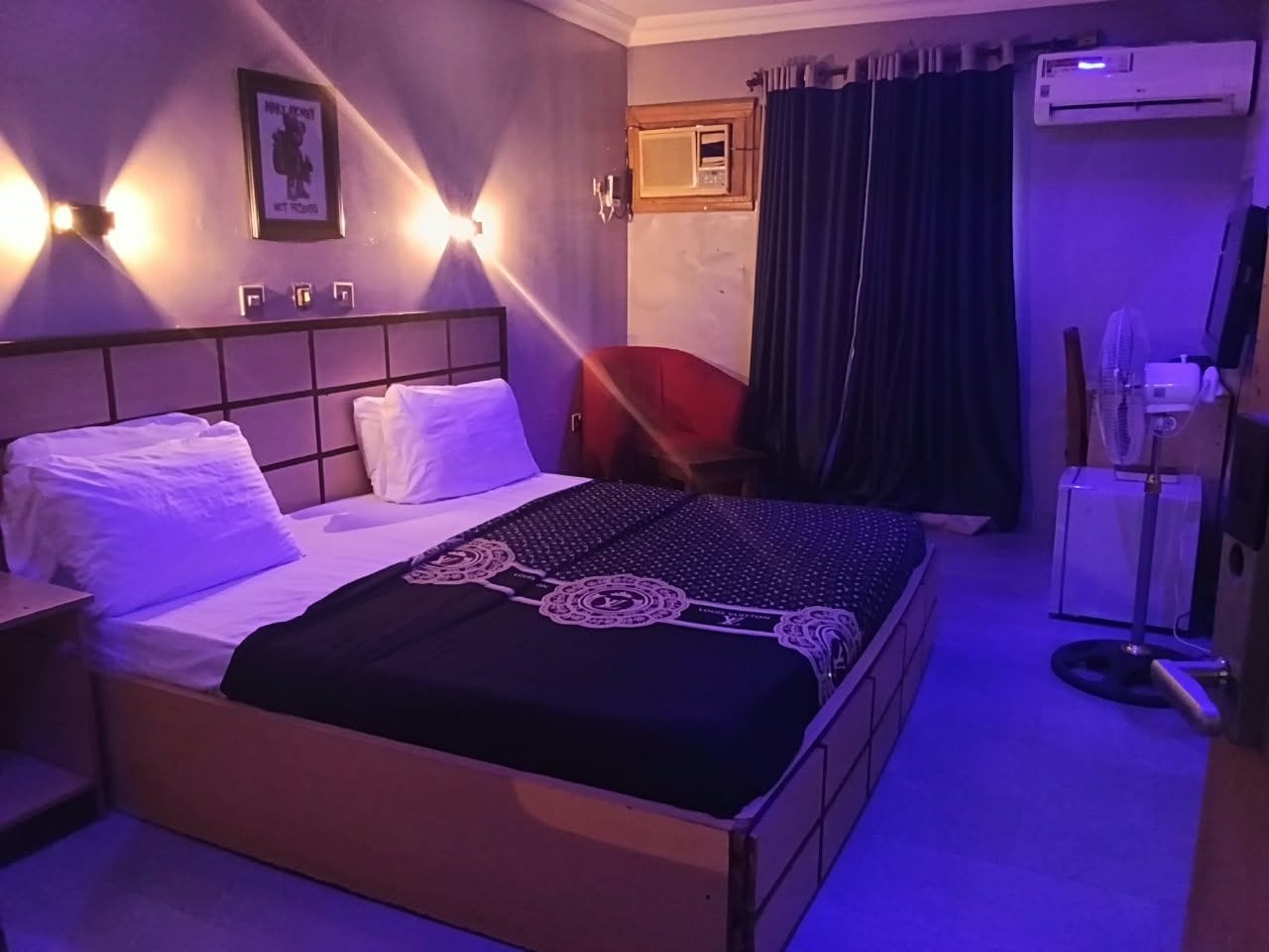 A hotel room with a bed, bedside table, wall-mounted light, air conditioning unit, and a window with curtains.