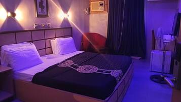 A hotel room with a bed, bedside table, wall-mounted light, air conditioning unit, and a window with curtains.