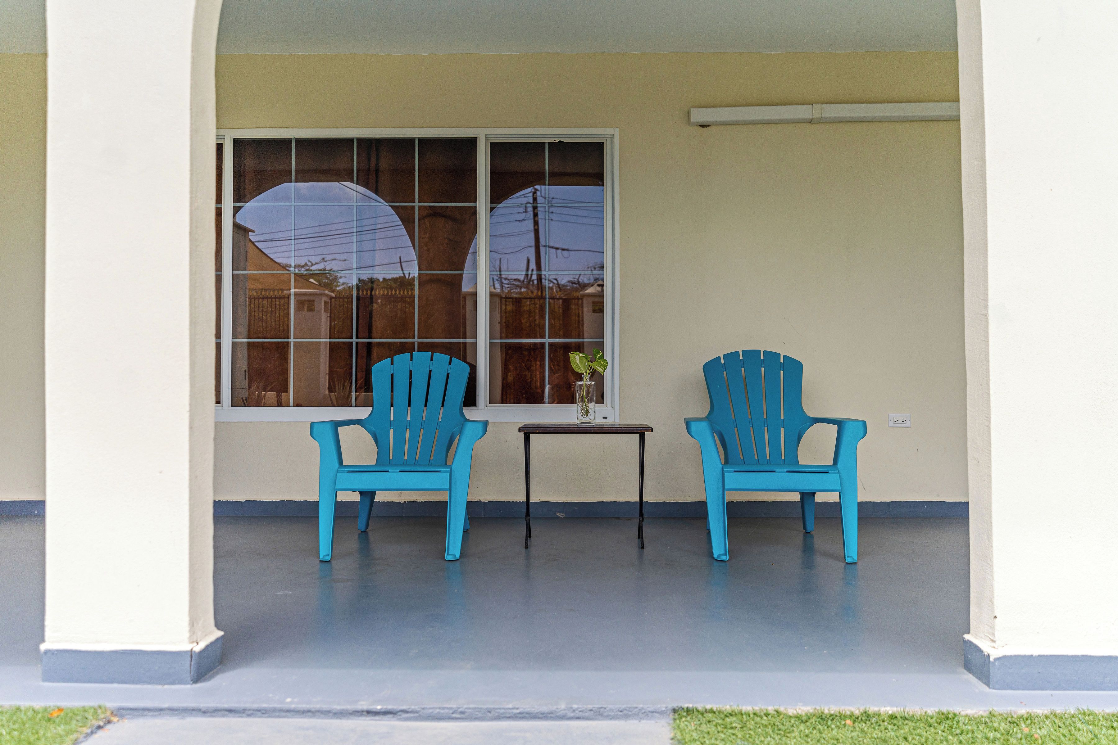 A covered porch with two blue chairs and a small table, a window with a view of a fence and trees, and a white wall.