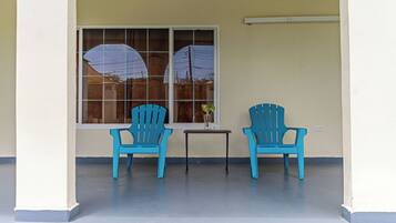 A covered porch with two blue chairs and a small table, a window with a view of a fence and trees, and a white wall.