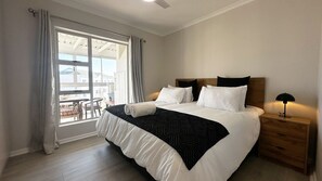 A hotel room with a large bed, a wooden headboard, a nightstand with a lamp, and a view of the outside through a window with curtains. - Perfect 2-bedroom apartment in Big Bay with stunning views (Cape Town)