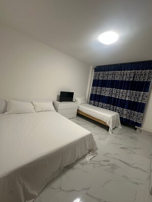 A bedroom with a large bed, a smaller bed, a bedside table with a TV, and a window with patterned curtains. - New Listing (Venezia)