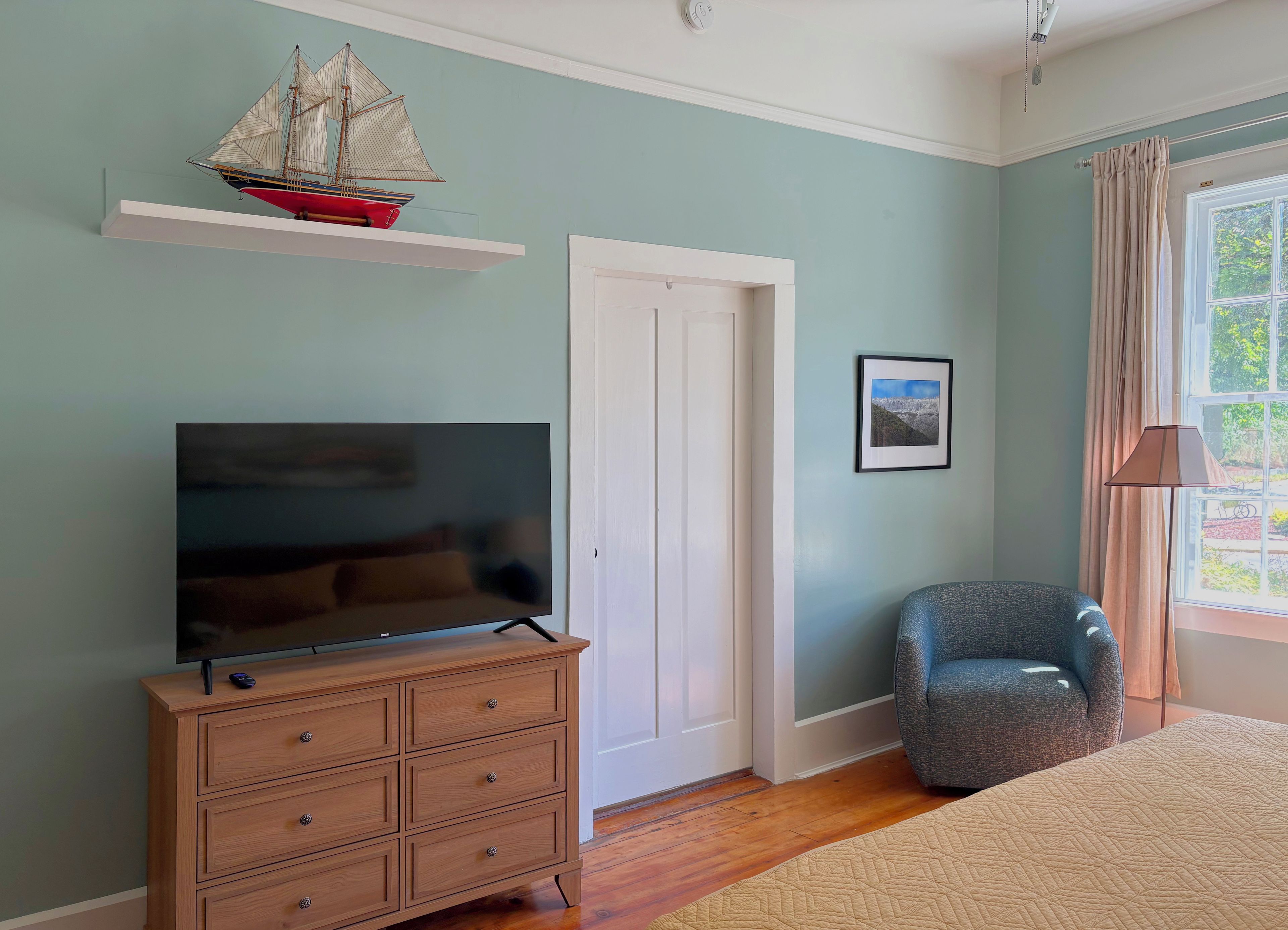 A bedroom with a television on a wooden dresser, a chair, a door, and a window with curtains.