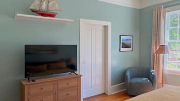 A bedroom with a television on a wooden dresser, a chair, a door, and a window with curtains.