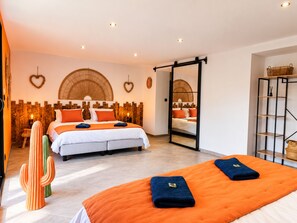 Ein Hotelzimmer mit einem großen Bett, einem kleineren Bett, einem Spiegel, einer Zimmerpflanze im Kaktus-Stil und einem Regal mit Körben.