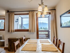 A dining area with a table set for four, wooden chairs, a view of mountains through large windows, and a chandelier. - Apartment with balcony with mountain view (Morillon)