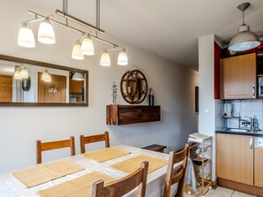 A dining area with a table set for four, wooden chairs, a hanging light fixture, a mirror, and a kitchen area with cabinets and appliances. - Apartment with balcony with mountain view (Morillon)