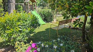 A playground with a slide and swings, surrounded by lush greenery and flowering plants.