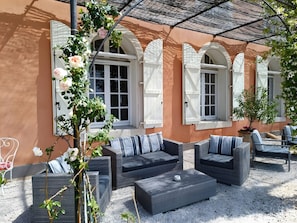 Un área de estar al aire libre con mobiliario de mimbre, una pérgola y un edificio con ventanas arqueadas.