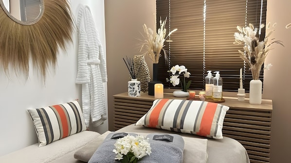 Un cuarto de spa con una camilla de masaje, almohadones a rayas, un florero con plantas secas, una vela y un espejo.