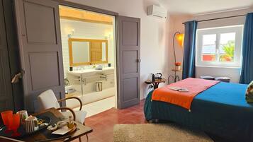 Habitació d'hotel amb un llit, una escriptori amb una safata d'objectes, un bany amb un lavabo i un mirall, i una finestra amb cortines.