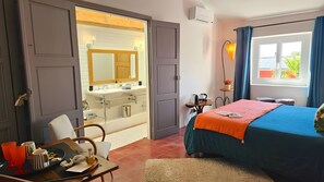 Habitación de hotel con una cama, un escritorio con una bandeja de objetos, un baño con lavamanos y espejo, y una ventana con cortinas.