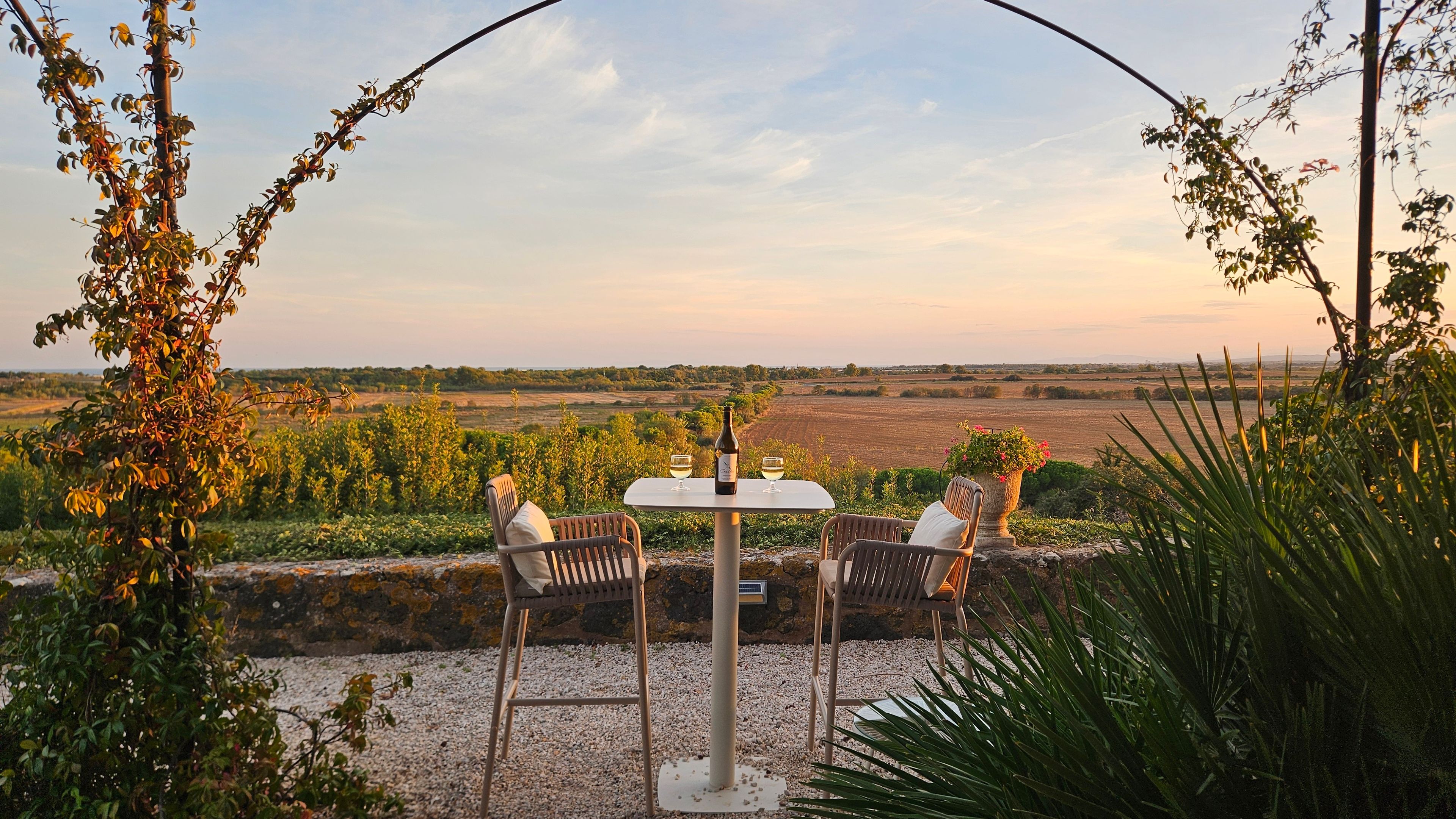 A table with two chairs and a bottle of wine, set against a scenic view of a field and distant horizon.