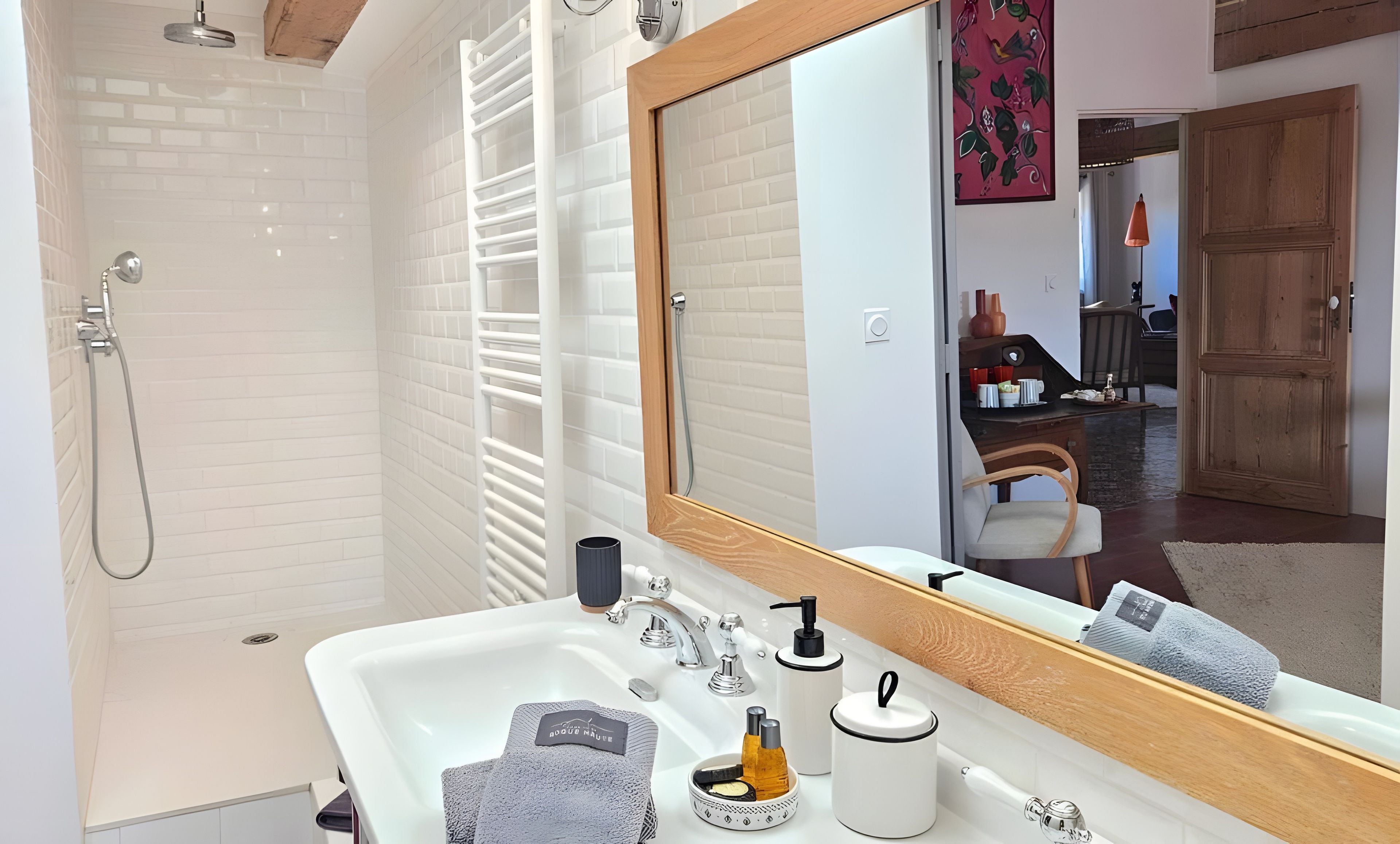 Een badkamer met een douche, een wastafel met toiletartikelen en een spiegel die een kamer met een deur en een stoel weerspiegelt.