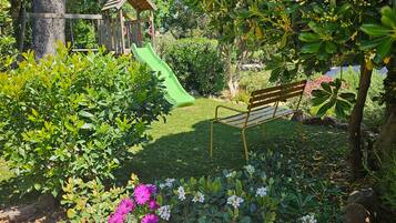 A playground with a slide and swings, surrounded by lush greenery and flowering plants.