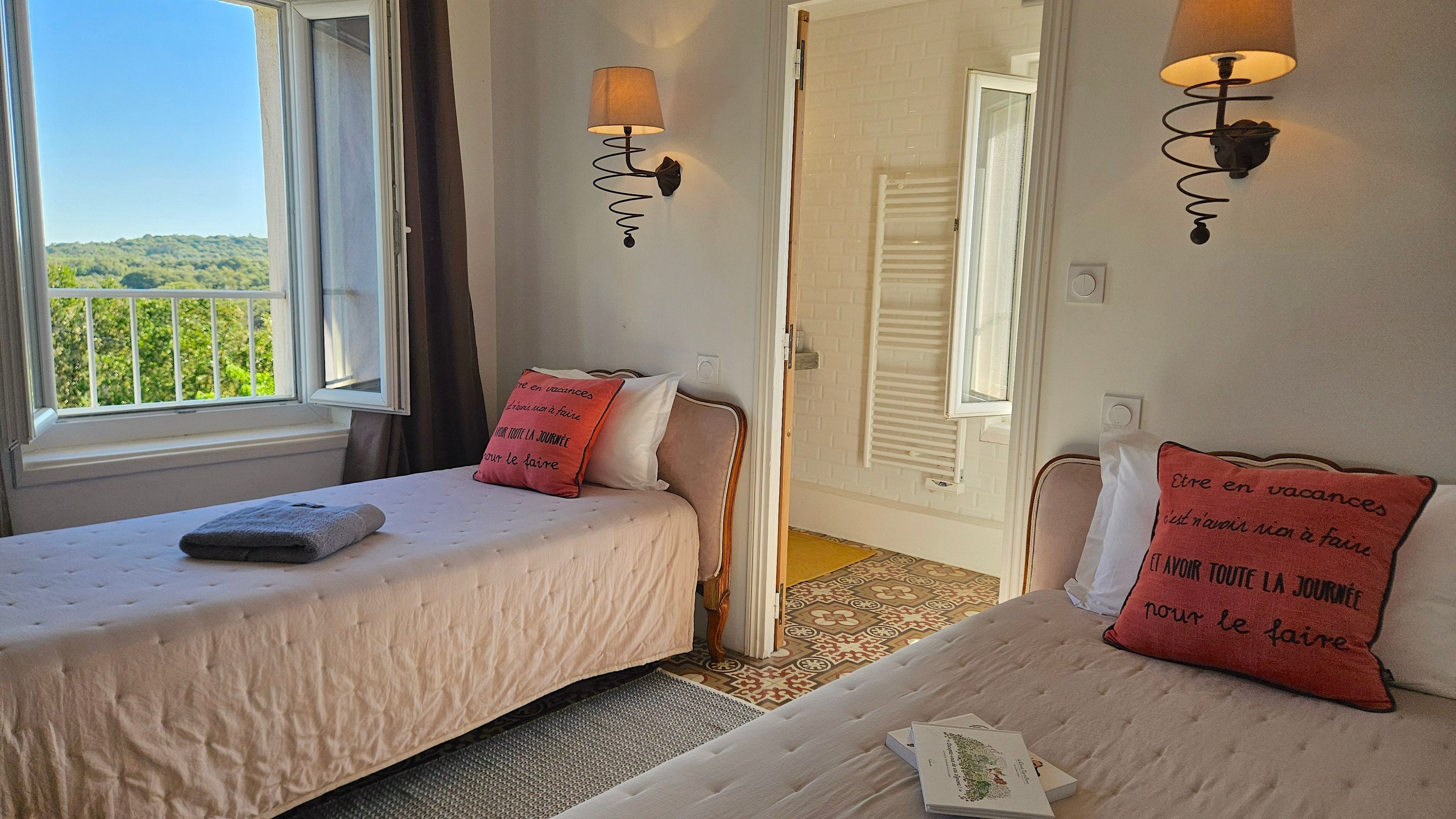 A hotel room with two beds, a window with a view of greenery, and wall-mounted lamps.