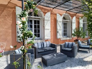 Ett utomhusområde med sittplatser, möbler i rotting, en pergola och en byggnad med välvda fönster.