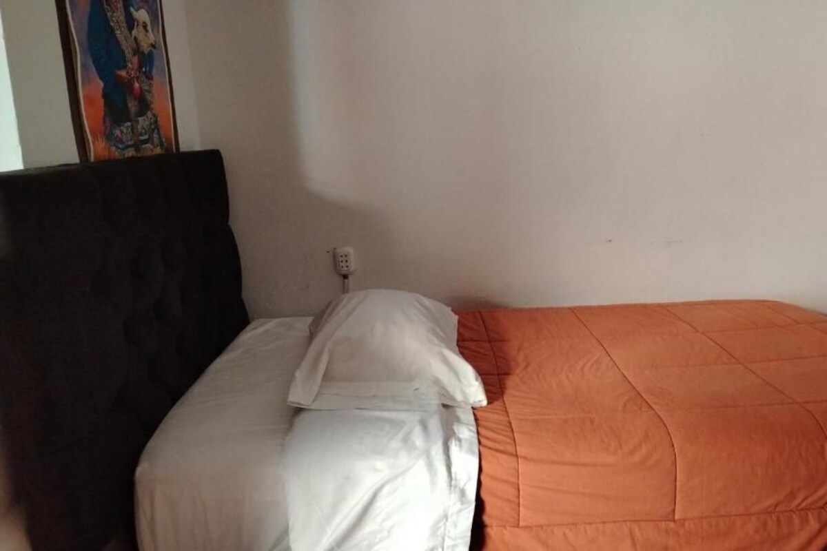 Un lit simple avec une couvre-lit blanche et orange, une tête de lit sombre et une prise de courant fixée au mur.