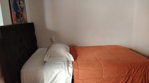 Una cama individual con colcha blanca y naranja, cabecera oscura y un enchufe en la pared.