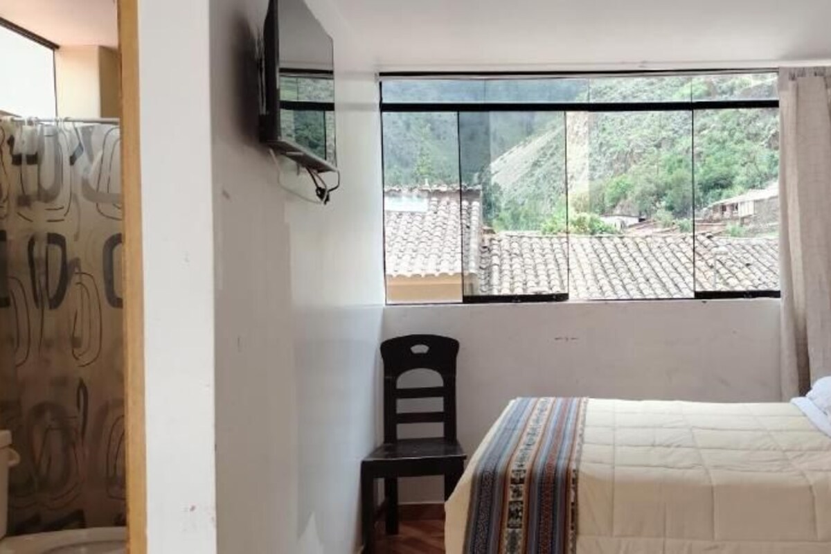 Une chambre à coucher avec un lit, une chaise, une fenêtre donnant sur un village et un téléviseur fixé au mur.