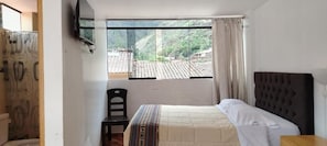 Un dormitorio con cama, una silla, ventana con vistas a un pueblo y televisor en la pared.
