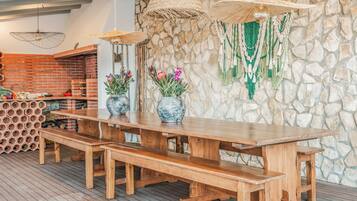 Una mesa de madera para comer con bancos, rodeada de paredes de piedra y decoraciones colgantes de macramé.