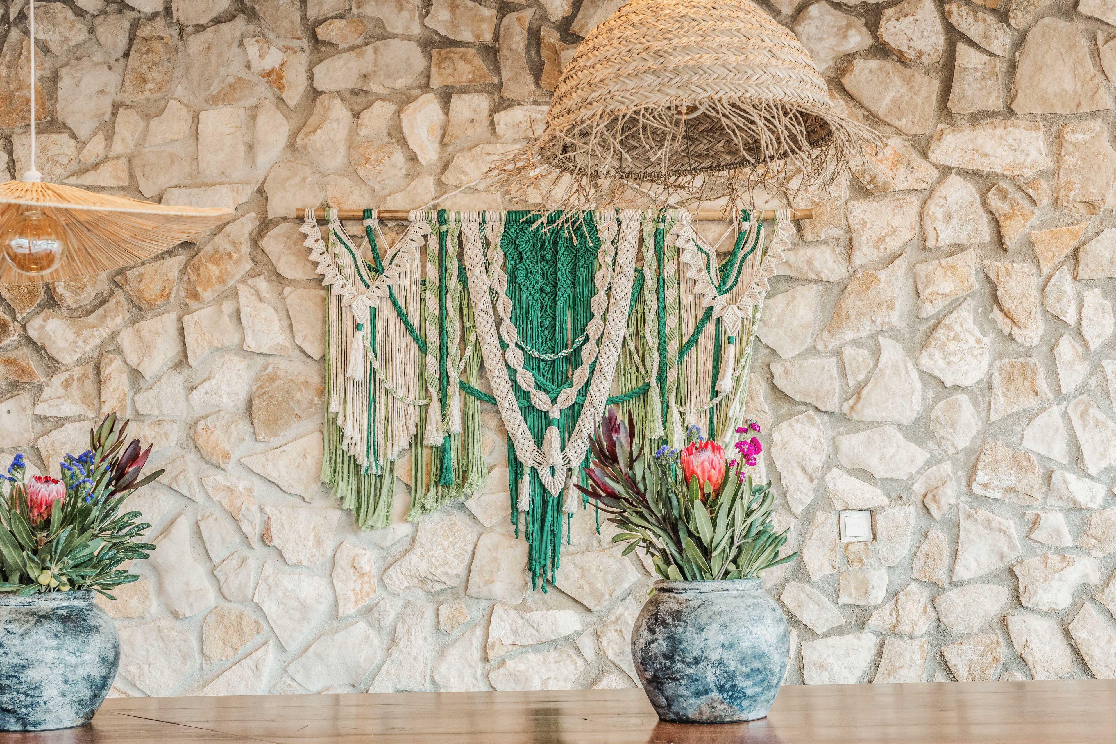 A stone wall with a macrame wall hanging and two potted plants with flowers.