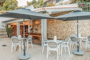 Une terrasse extérieure avec des chaises et des tables blanches, des murs en pierre et des parasols.