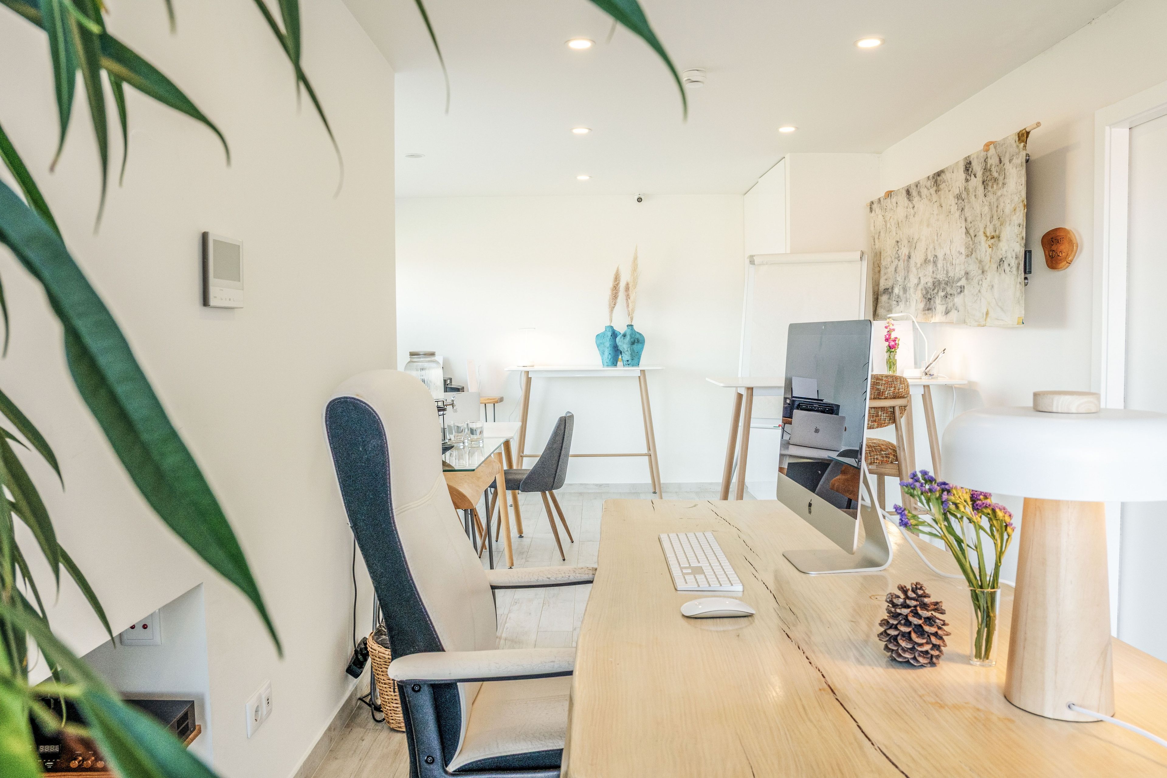 A modern office space with a wooden desk, a computer, and a chair. There is a whiteboard and a vase with flowers on the wall.