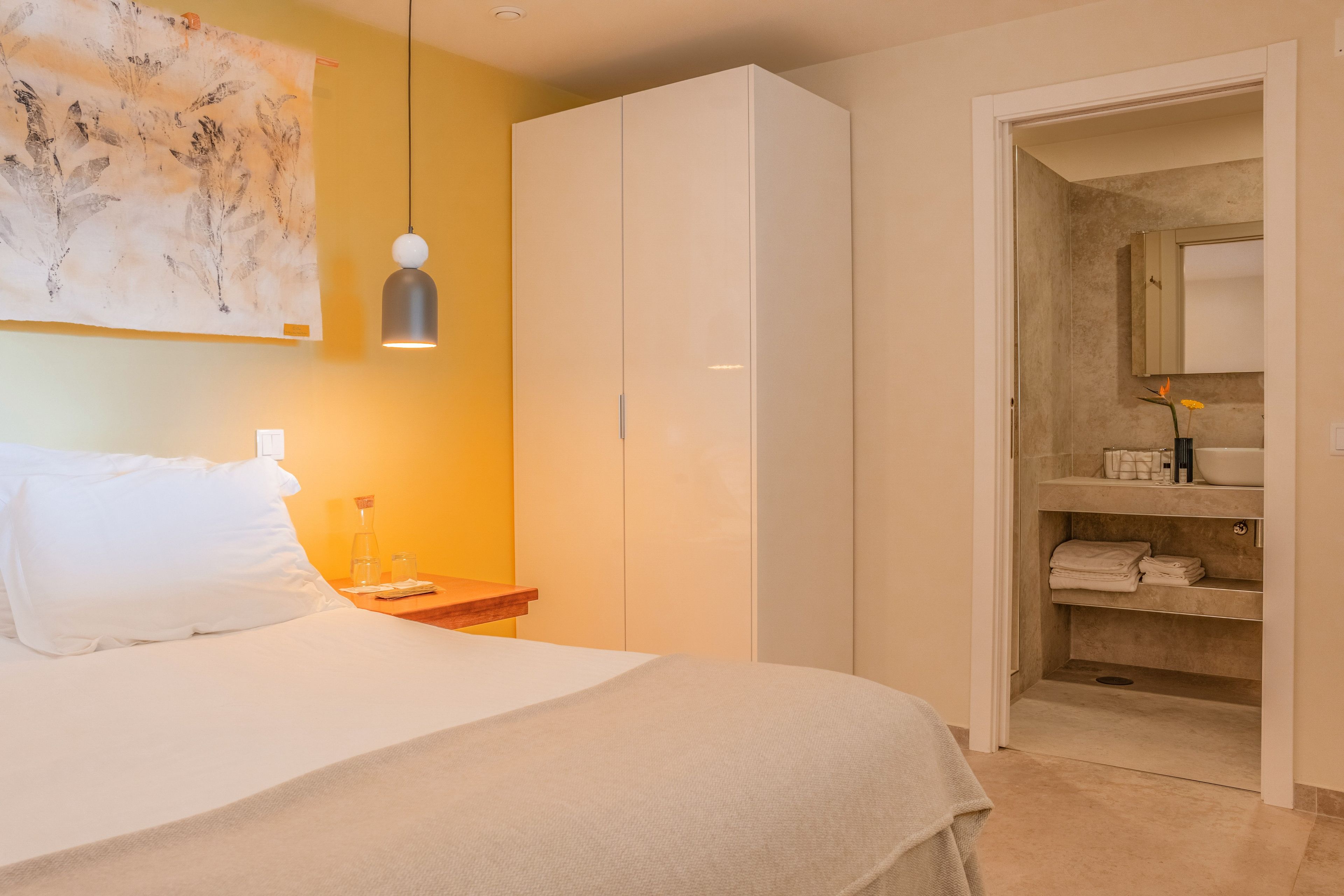 Ein Hotelzimmer mit Bett, Nachttisch mit Lampe, Kleiderschrank und Bad mit Handtüchern und Toilettenartikeln.