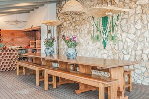 Une table à manger en bois, avec des bancs, entourée de murs en pierre et agrémentée de décorations en macramé suspendues.
