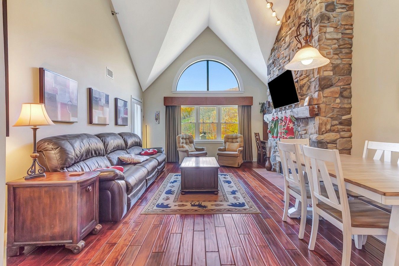 A spacious living room with a high ceiling, a stone fireplace, a dining area, and a large window providing natural light.