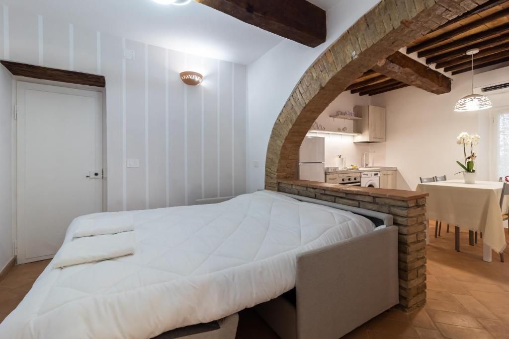 A bedroom with a bed, a stone archway, a kitchen in the background, and a dining area with a table and chairs.