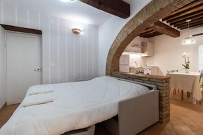 Un dormitori amb un llit, una arcada de pedra, una cuina al fons i una zona de menjador amb taula i cadires.