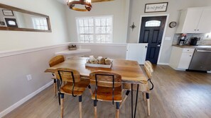 Un coin repas avec une table et des chaises en bois, une cuisine avec des meubles blancs et une porte sur laquelle est affiché un panneau indiquant : « Soyez les bienvenus ».