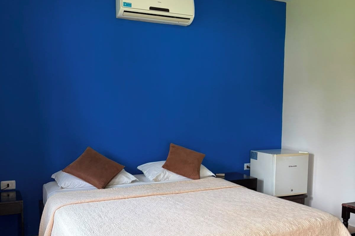 Una habitación de hotel con una cama, una mesita de noche, un equipo de aire acondicionado y una pequeña mesa con lámpara.