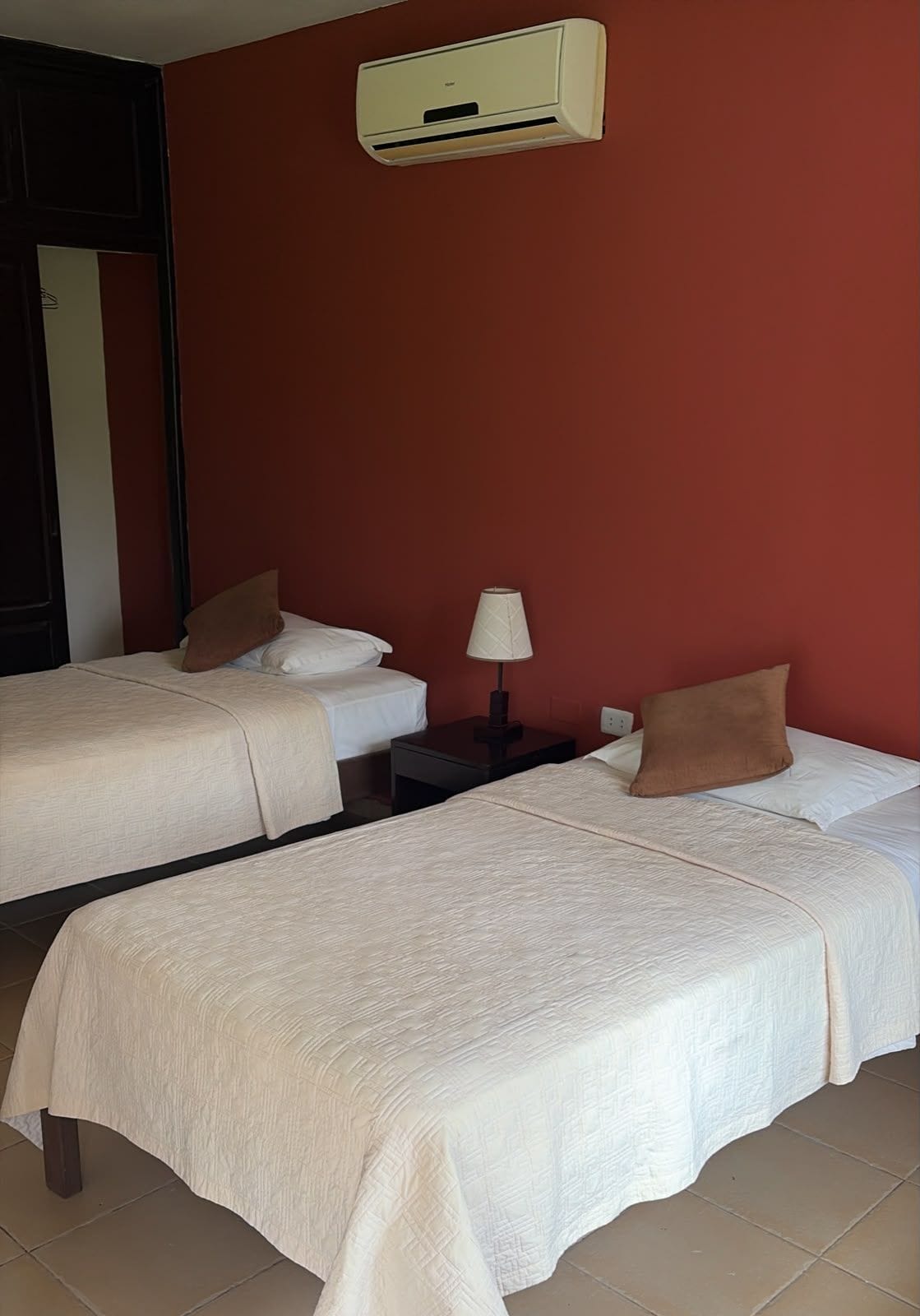 A hotel room with two beds, a nightstand with a lamp, and an air conditioning unit on the wall.