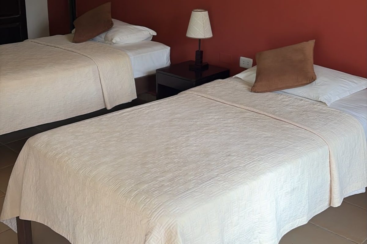 Una habitación de hotel con dos camas, una mesita de noche con lámpara y un equipo de aire acondicionado en la pared.