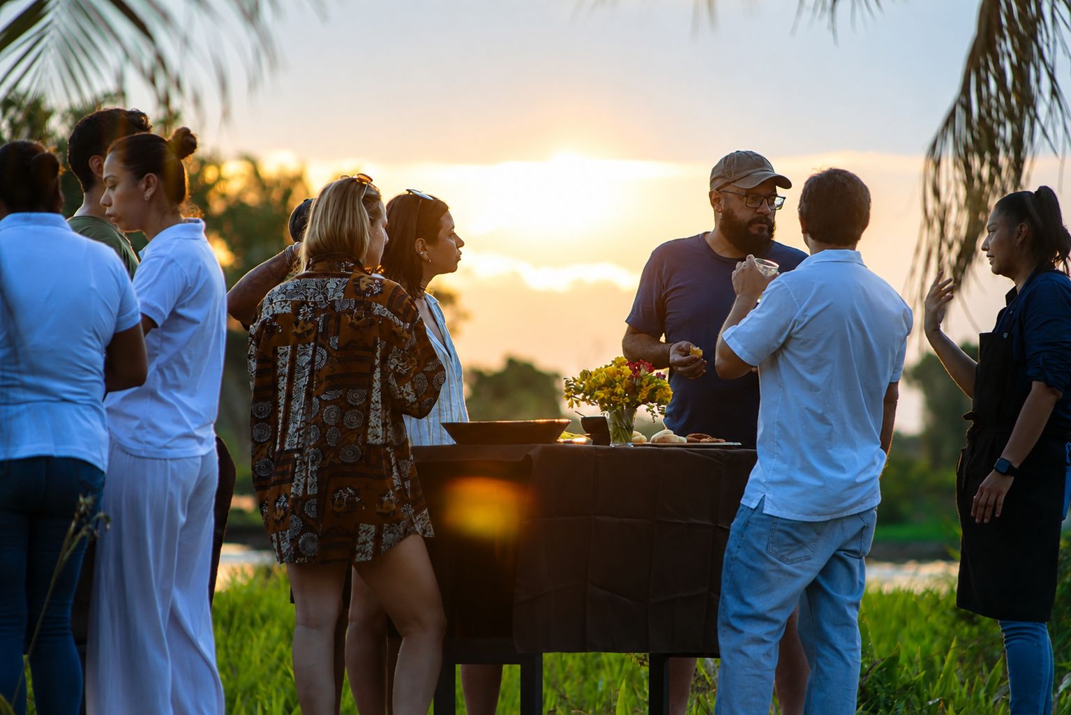 A group of people gathered around a table with food and drinks, outdoors during sunset.