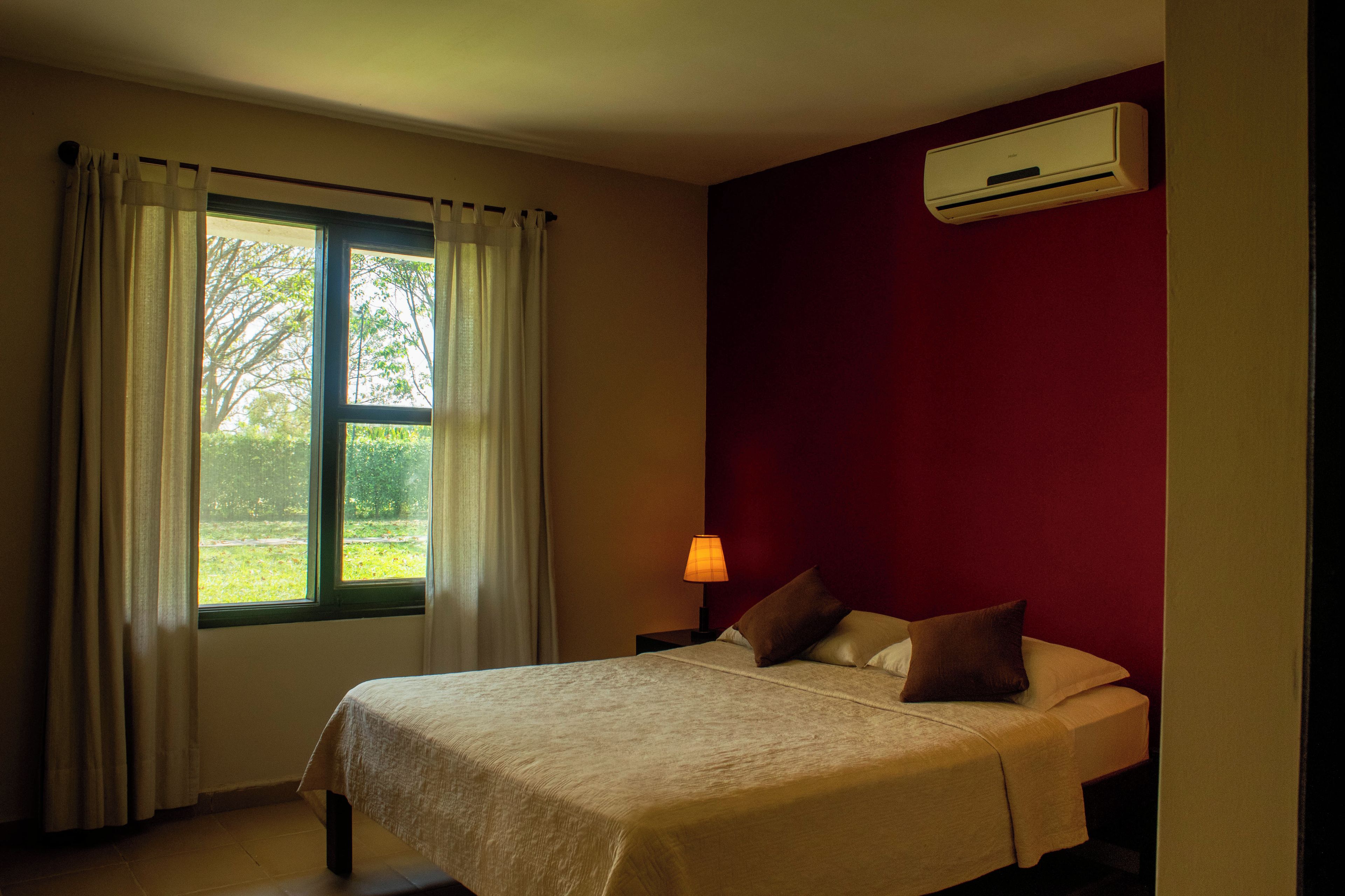 A bedroom with a bed, a window with curtains, a nightstand with a lamp, and an air conditioning unit on the wall.