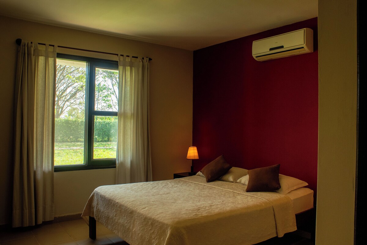 Un dormitorio con cama, ventana con cortinas, mesita de noche con lámpara y un equipo de aire acondicionado en la pared.