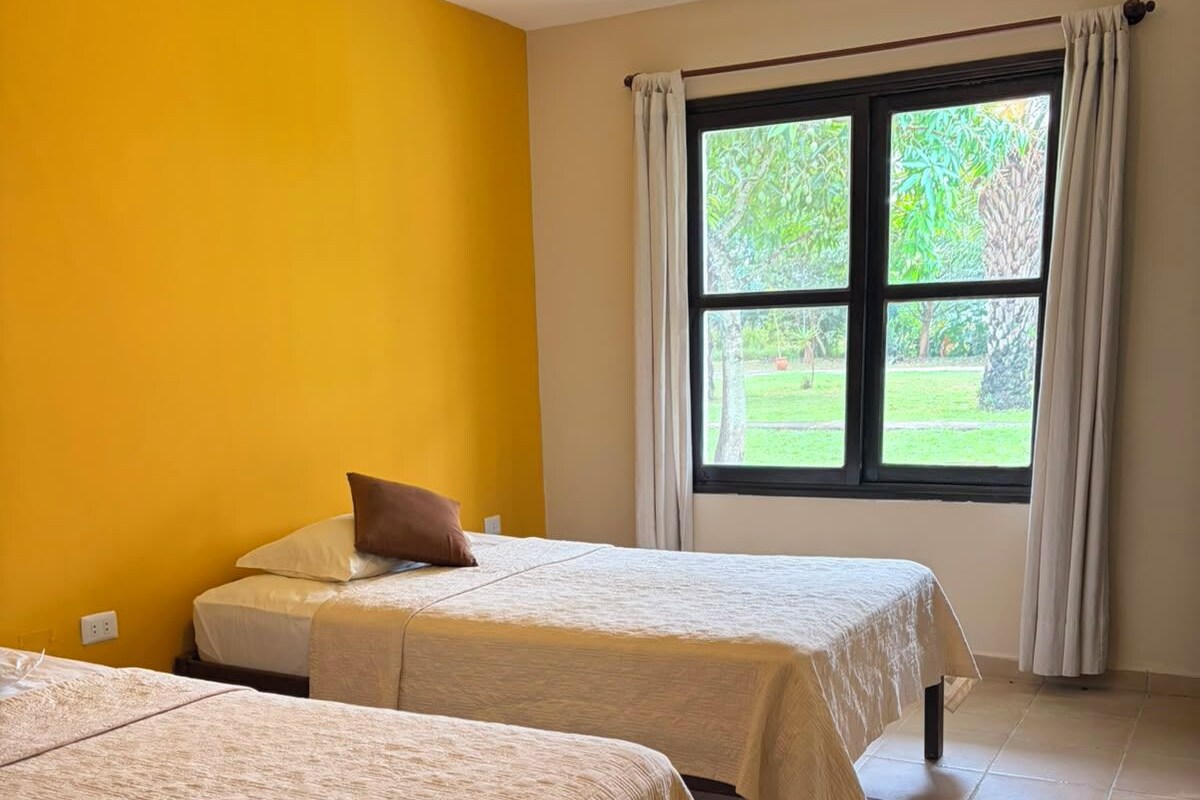 Una habitación con dos camas, una ventana con cortinas y una pared amarilla.
